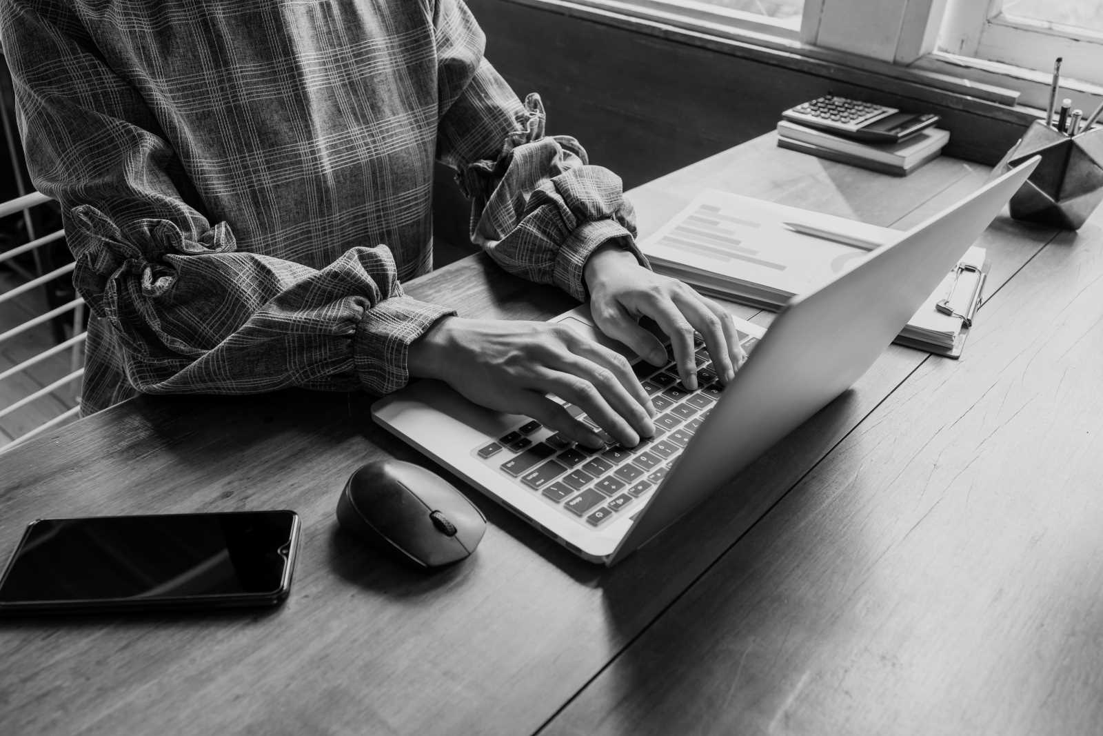 Person tippt am Laptop an einem Schreibtisch, mit Unterlagen, Smartphone und Maus – Fokus auf embedded software.